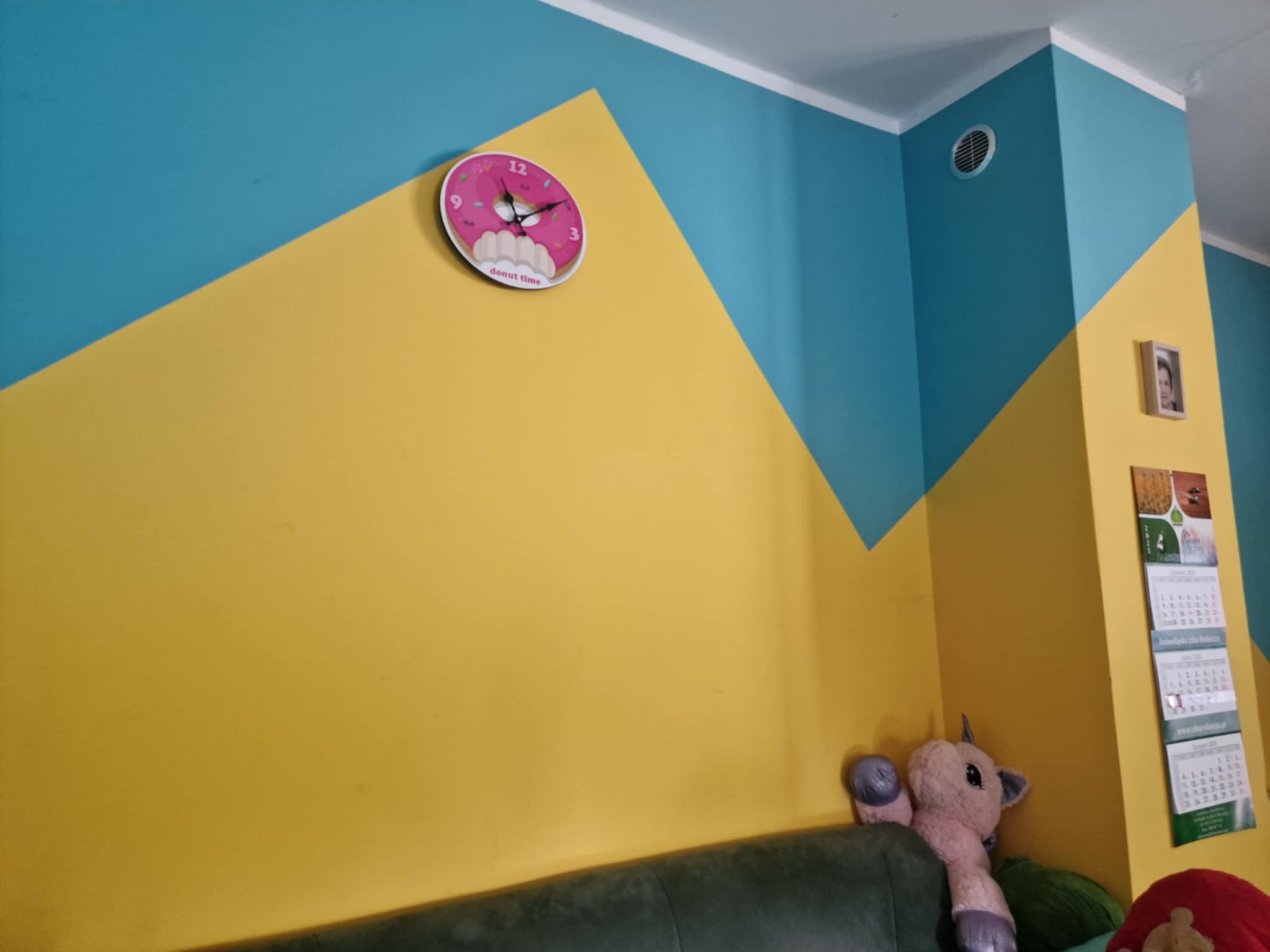 Pomysłowe malowanie ścian w pokoju dziecka: żółte i turkusowe trójkąty. Zegar w kształcie pączka, kalendarz i pluszowy jednorożec dodają uroku.