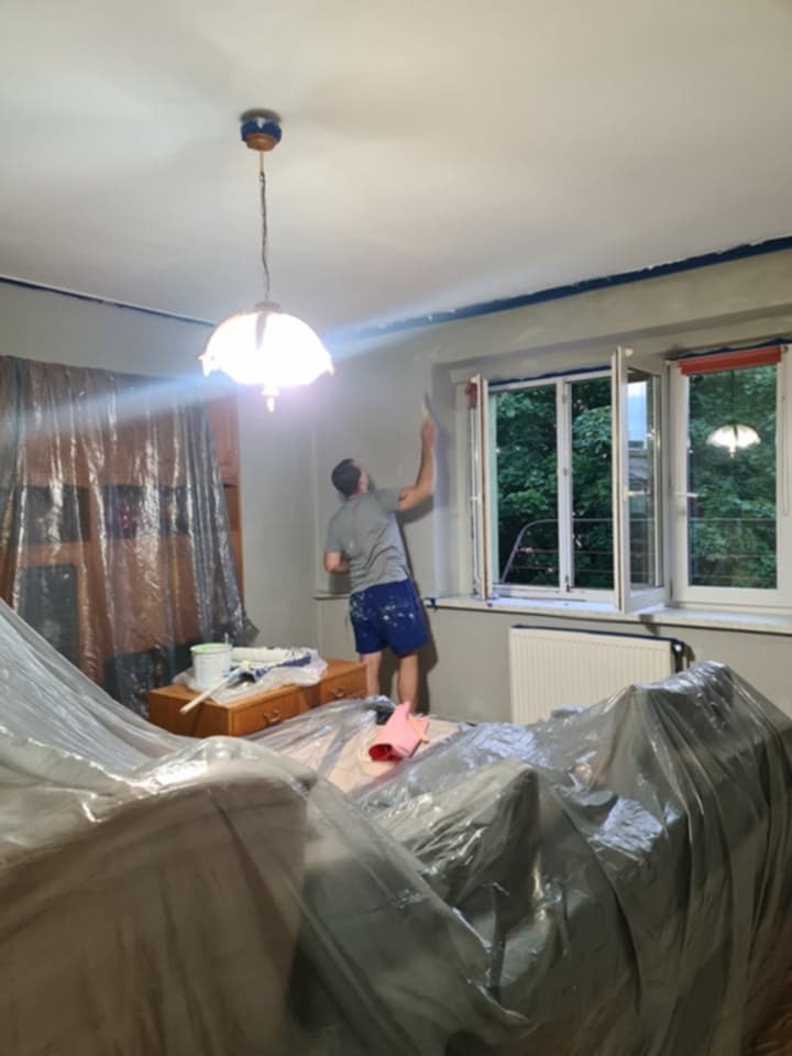 Malowanie ścian w pokoju z zabezpieczonymi meblami folią. Widoczny pracownik z wałkiem malarskim przy oknie i wiadro z farbą na komodzie.