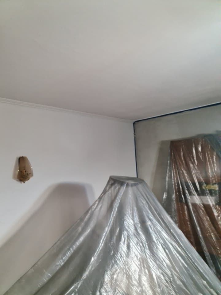 Pokój przygotowany do malowania: ściany z taśmą malarską, meble zakryte folią ochronną. Widoczny fragment sufitu i lampa na ścianie.