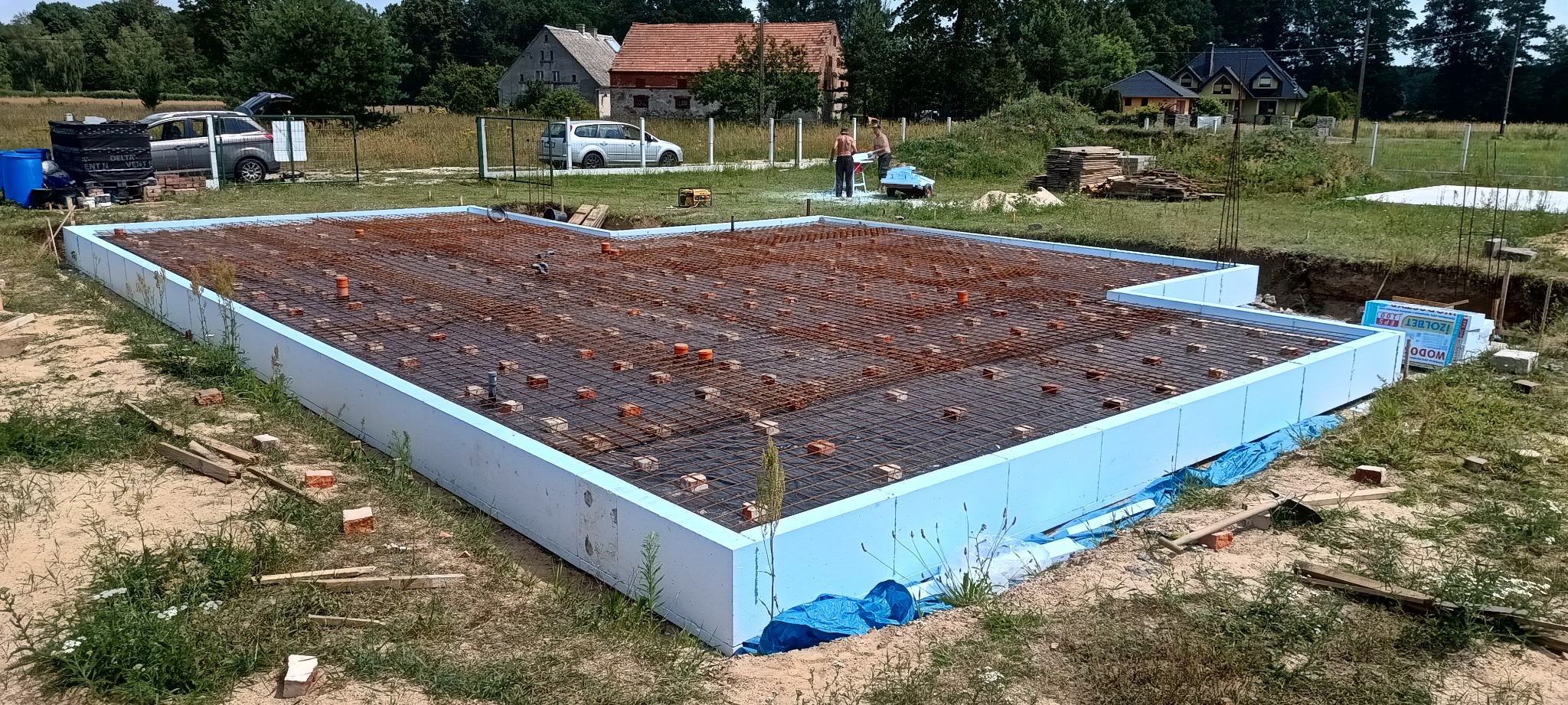 Zbrojenie fundamentów domu jednorodzinnego, otoczone niebieskim styropianem. W tle widoczni robotnicy i zabudowa jednorodzinna. Prace budowlane w toku.
