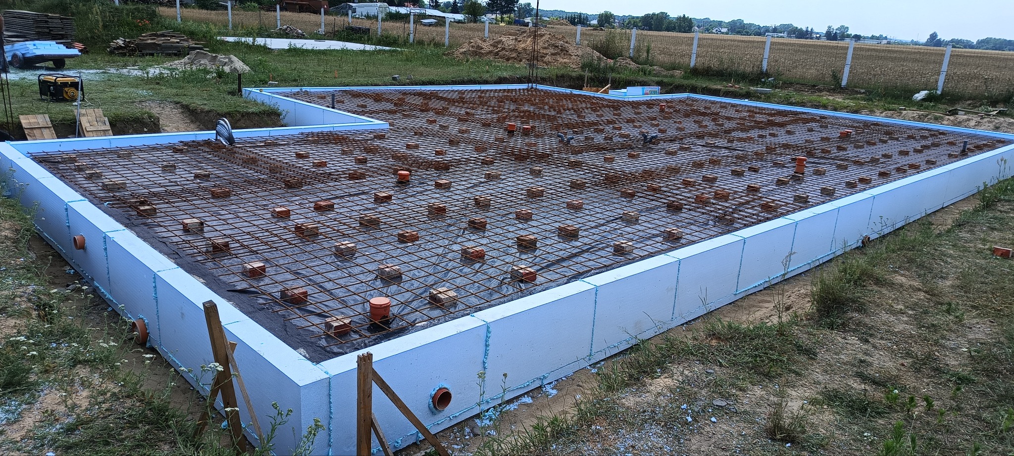 Zbrojenie fundamentów z izolacją pionową, widoczne cegły dystansowe, rury kanalizacyjne. Fundament zabezpieczony folią, w tle pole i ogrodzenie.