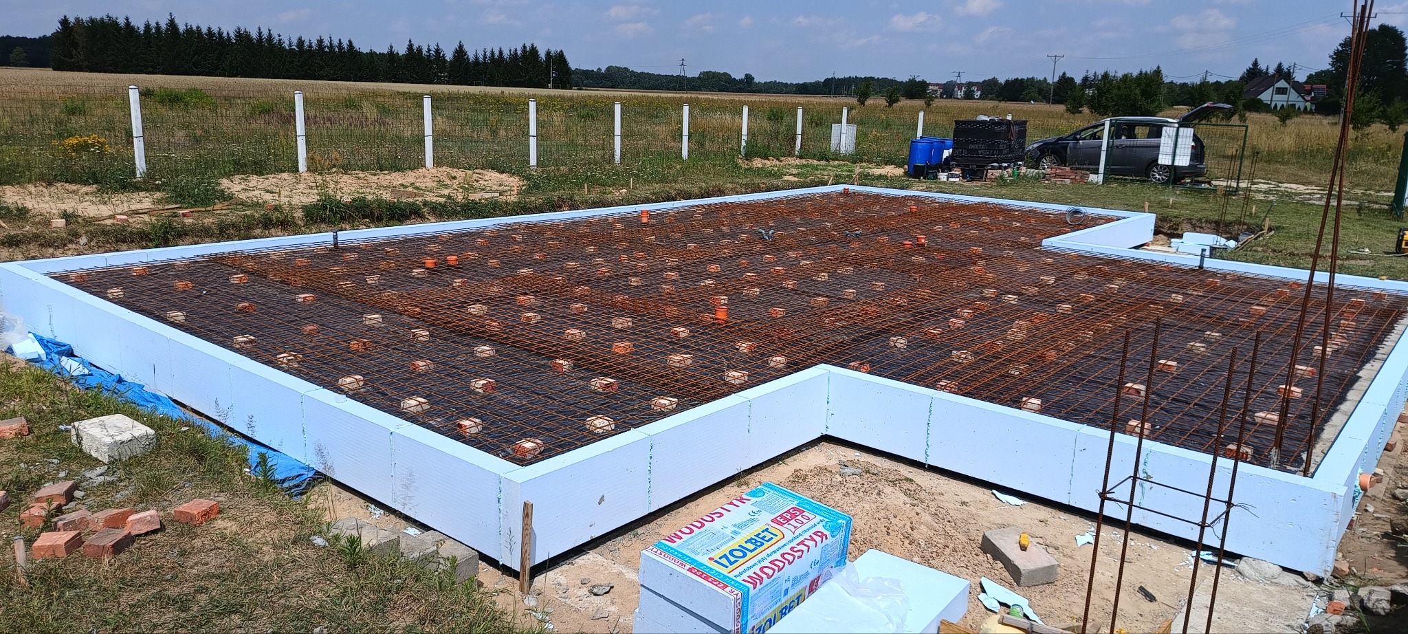 Fundamenty domu jednorodzinnego: szalunek z izolacją, zbrojenie siatką i kostkami dystansowymi. Widok z góry na plac budowy w słoneczny dzień.