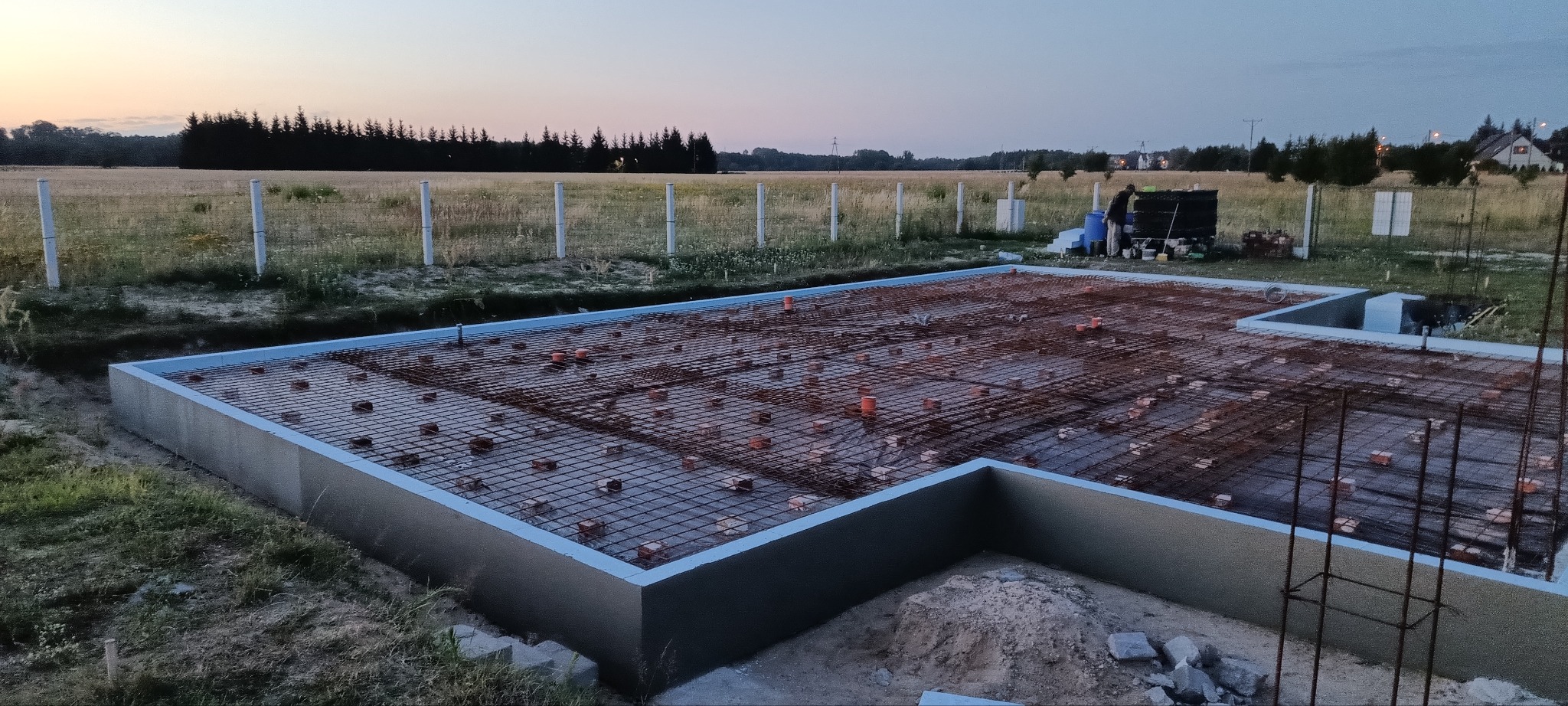 Fundamenty domu jednorodzinnego zbrojone stalą, widoczne elementy instalacji, otoczone polami i lasem o zmierzchu.