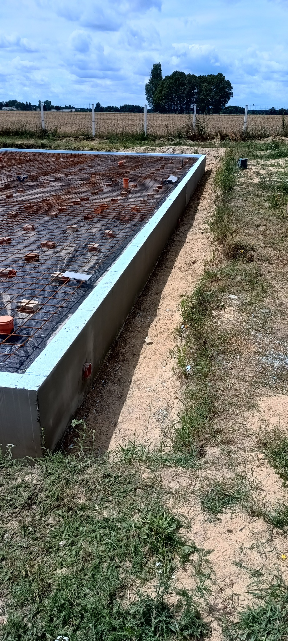 Zbrojenie fundamentów z siatką, cegłami i rurami PCV, przygotowane do zalania betonem. Widok z boku na teren budowy w słoneczny dzień.