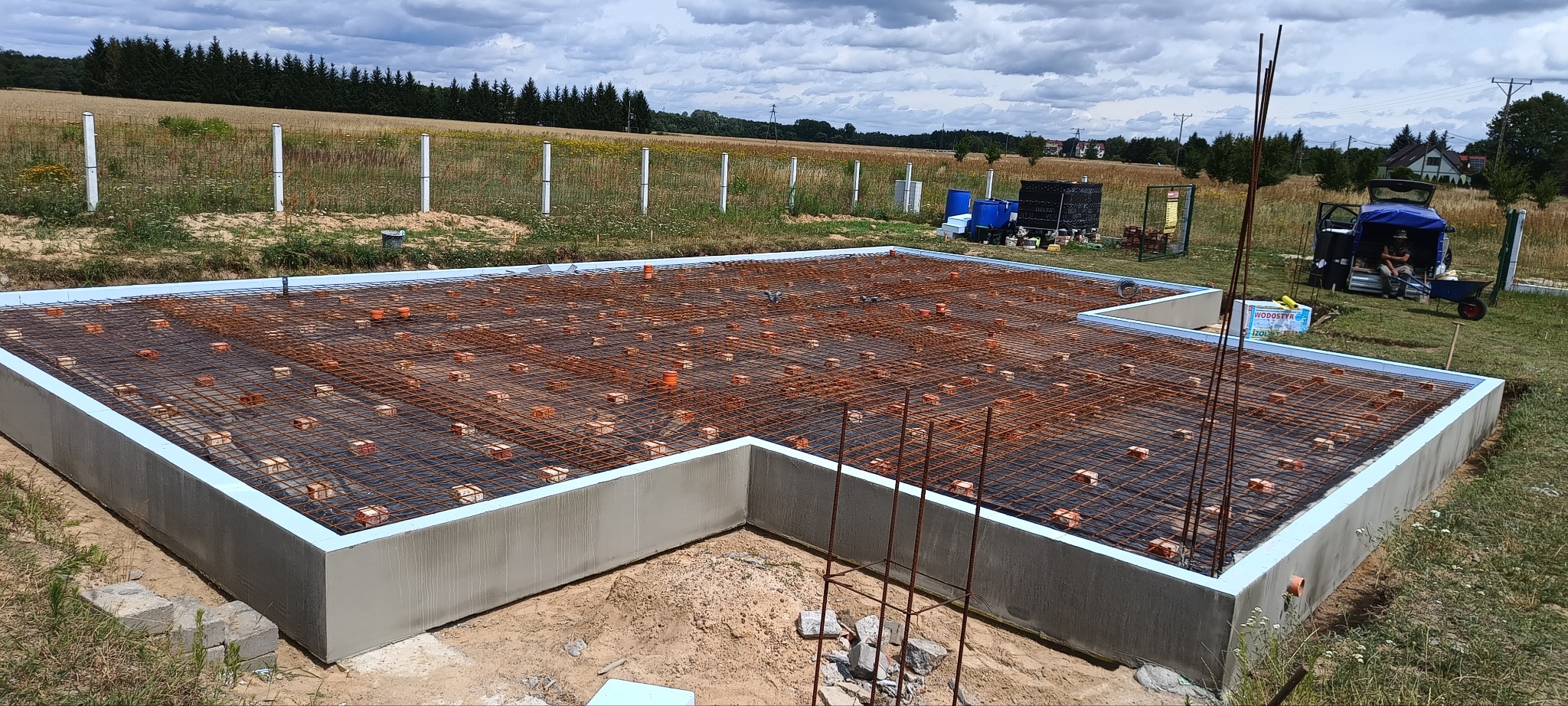 Fundament domu w trakcie budowy: zbrojenie, izolacja termiczna, elementy instalacji. Widok z góry na przygotowany szkielet konstrukcji na tle krajobrazu.