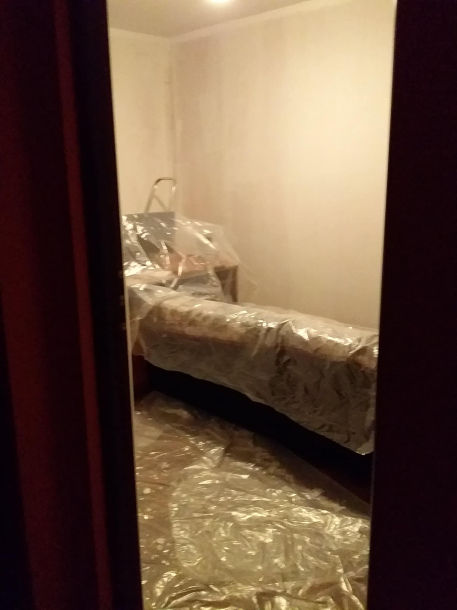 Pokój przygotowany do malowania: meble zabezpieczone folią, drabina w rogu. Widok zza framugi.