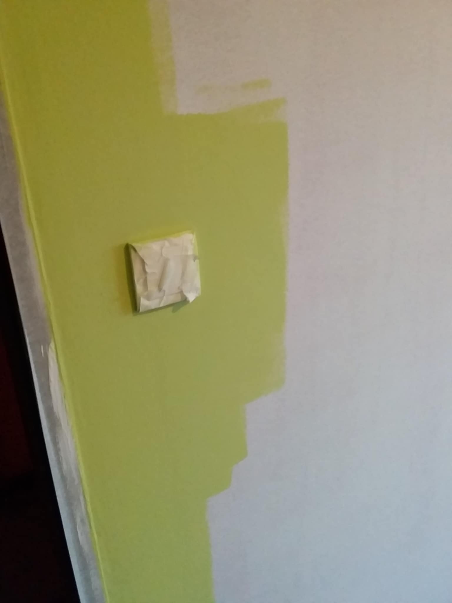 Fragment ściany w trakcie malowania: świeża, zielona farba kontrastuje z białym obszarem. Widoczna taśma malarska zabezpieczająca włącznik.