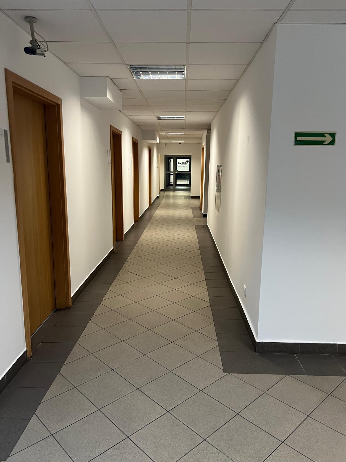 Długi korytarz biurowy z białymi ścianami, drewnianymi drzwiami i szarą podłogą w romby. Widoczny znak ewakuacyjny i kamera przemysłowa.