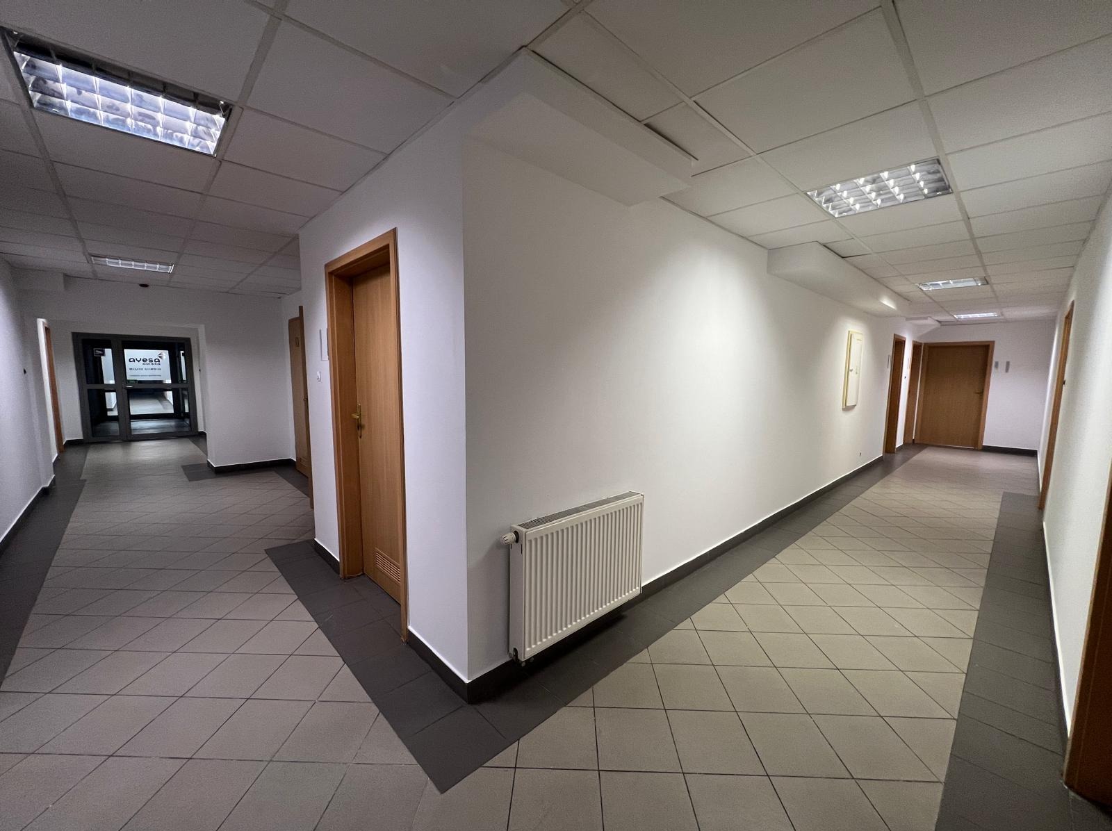 Korytarz biurowy z białymi ścianami, grzejnikiem i drewnianymi drzwiami. Podłoga wyłożona płytkami w odcieniach szarości. Widoczne oświetlenie sufitowe.