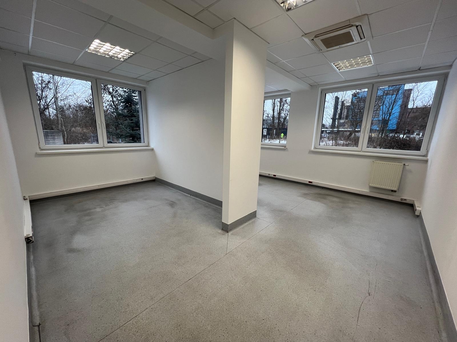Jasne, puste biuro po remoncie. Świeżo pomalowane ściany, okna z widokiem na zewnątrz, szara podłoga. Widoczny grzejnik i kolumna konstrukcyjna.