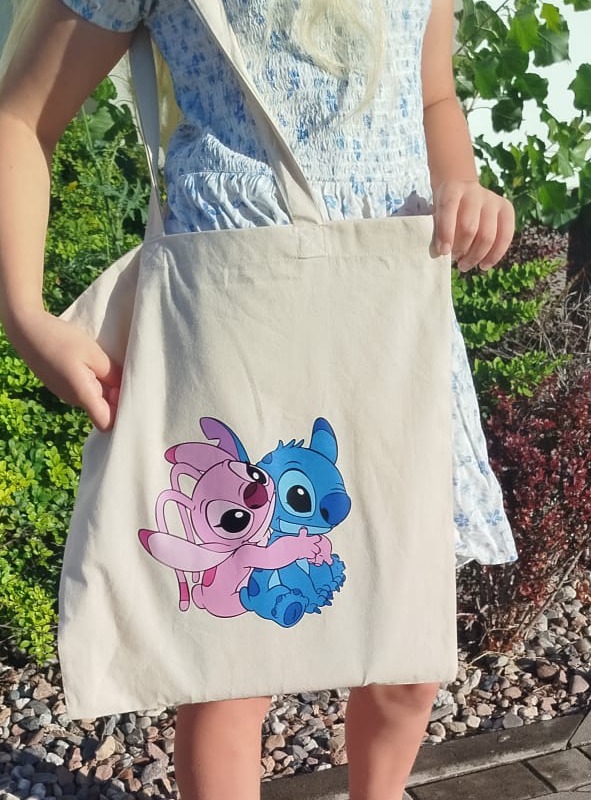 Dziewczynka prezentuje beżową torbę na ramię z nadrukiem postaci z bajki, trzymając ją za krawędzie. W tle widać zieloną roślinność.