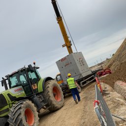 Oskar Kwaśny - Prace ziemne: Traktor CLAAS i dźwig transportują betonowy element H-SA1 Matbet na placu budowy. Pracownik w kasku i kamizelce nadzoruje proces.