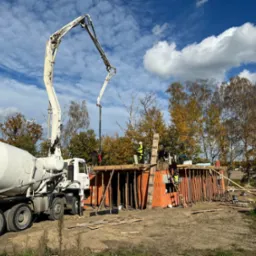 Betoniarka z pompą wylewa beton na fundament budynku z czerwonej cegły. Pracownicy w żółtych kamizelkach nadzorują proces na tle błękitnego nieba.