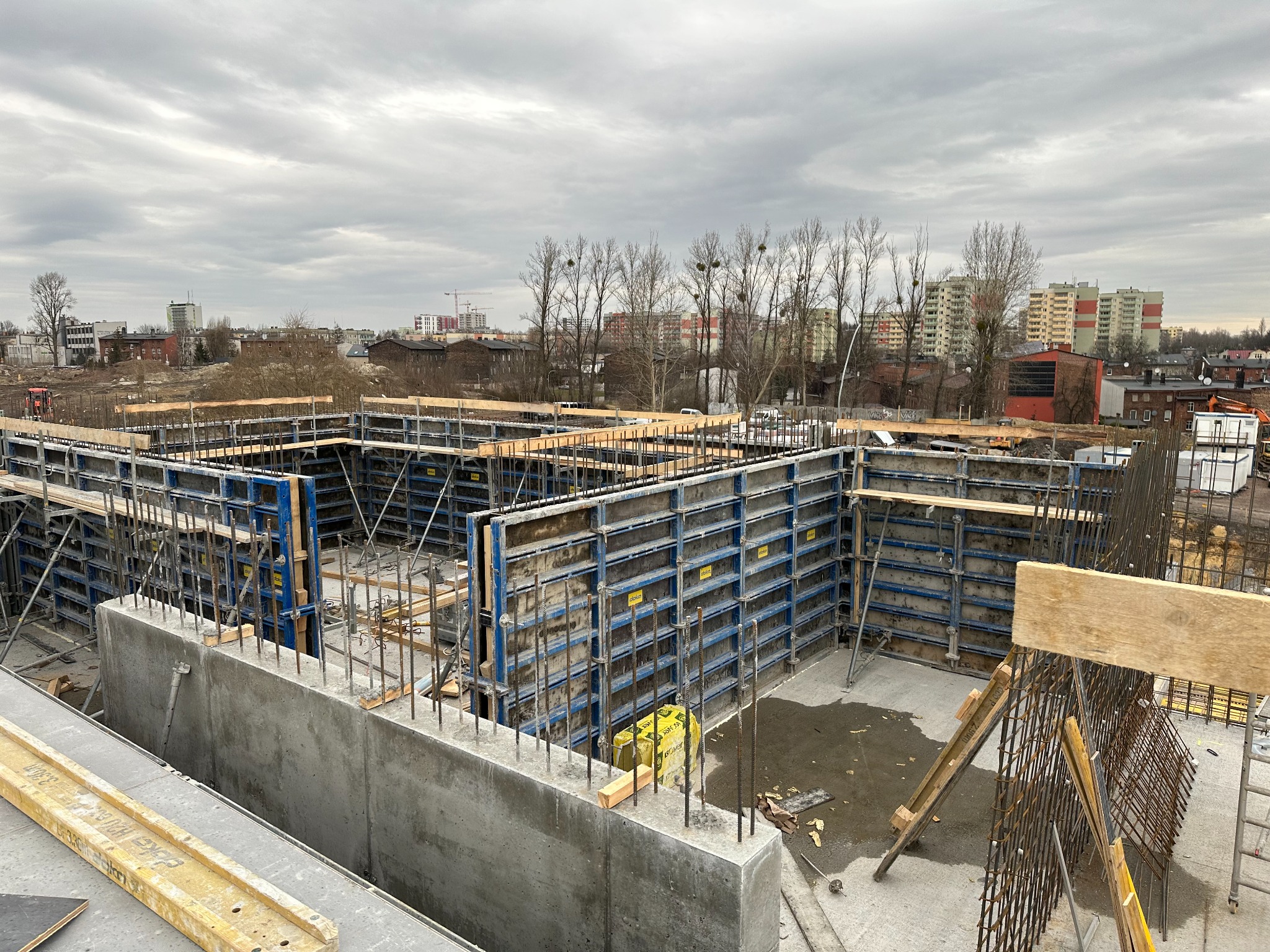 Wysoki kąt ujęcia fundamentów budynku w trakcie budowy, z widocznym zbrojeniem, szalunkami i drewnianymi elementami konstrukcyjnymi na tle miejskiego krajobrazu pod pochmurnym niebem.