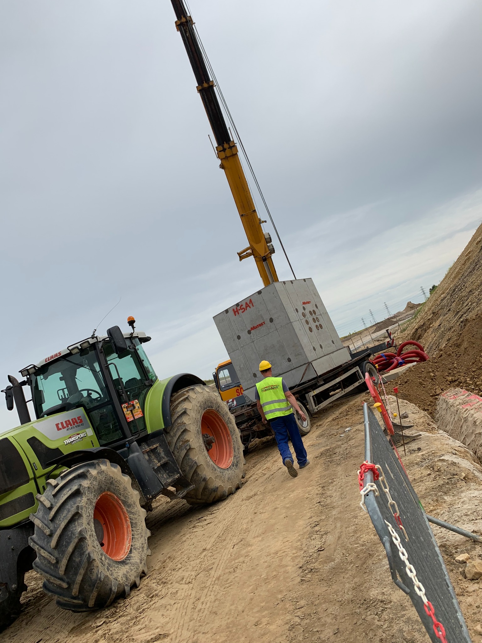 Prace ziemne: Traktor CLAAS i dźwig transportują betonowy element H-SA1 Matbet na placu budowy. Pracownik w kasku i kamizelce nadzoruje proces.