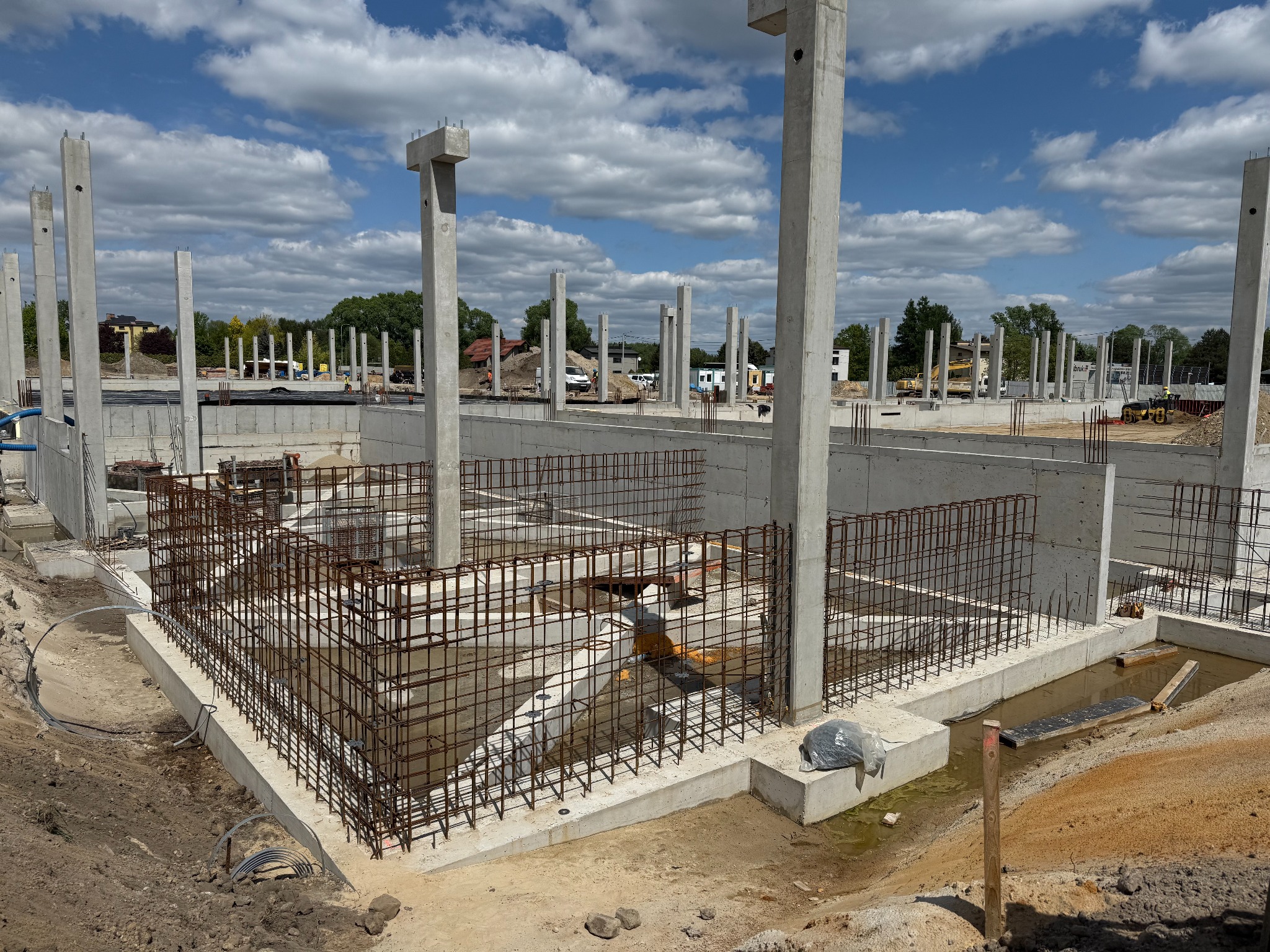 Budowa fundamentów z żelbetowymi słupami i zbrojeniami, widok na plac budowy z maszynami w tle, błękitne niebo z chmurami.