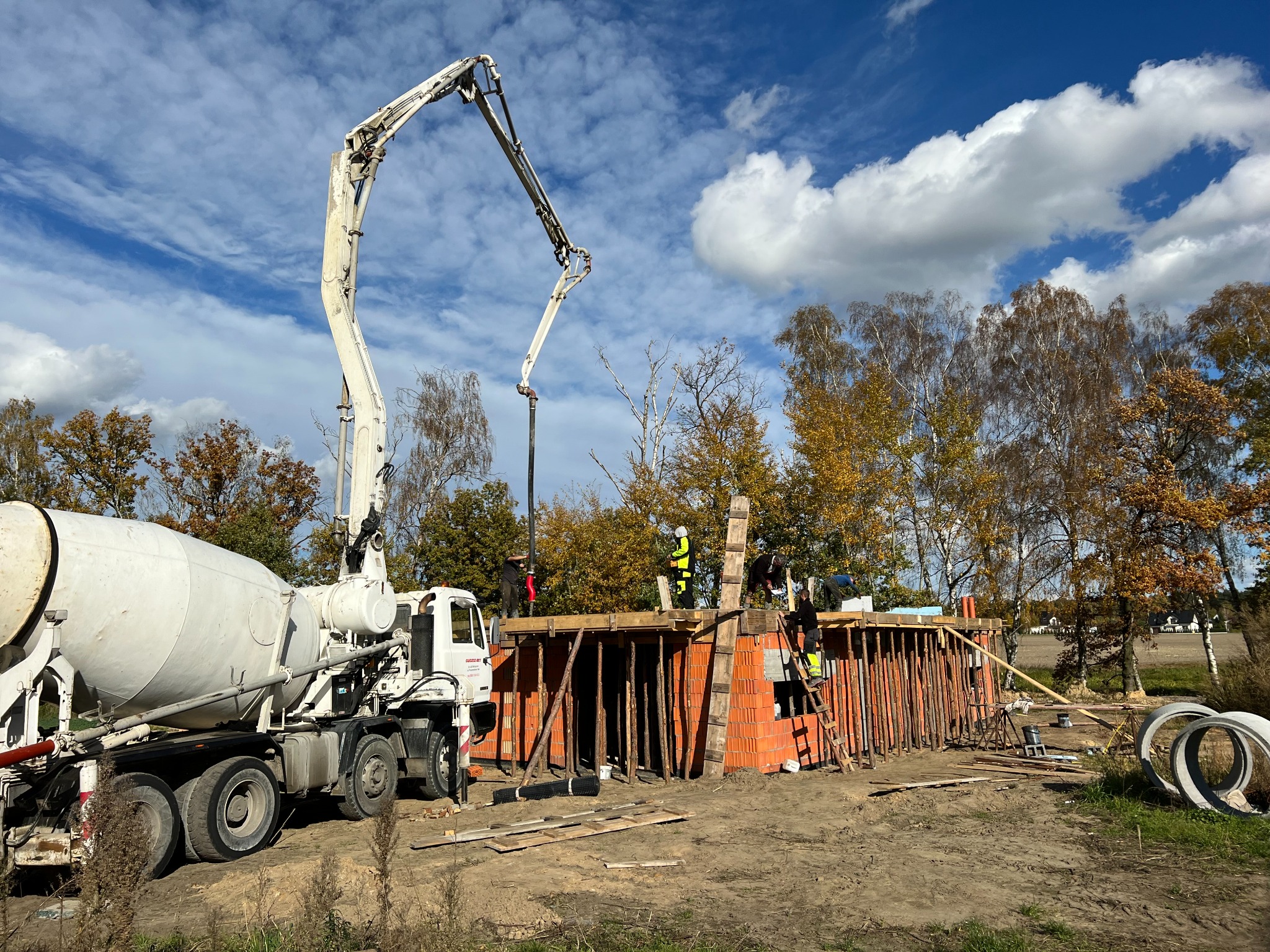 Betoniarka z pompą wylewa beton na fundament budynku z czerwonej cegły. Pracownicy w żółtych kamizelkach nadzorują proces na tle błękitnego nieba.