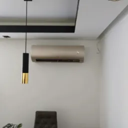 Klimatyzator w jasnym wnętrzu z lampą wiszącą. Widoczne elementy dekoracyjne sufitu. Minimalistyczny design, nowoczesne wykończenie. Kabel wystający ze ściany.