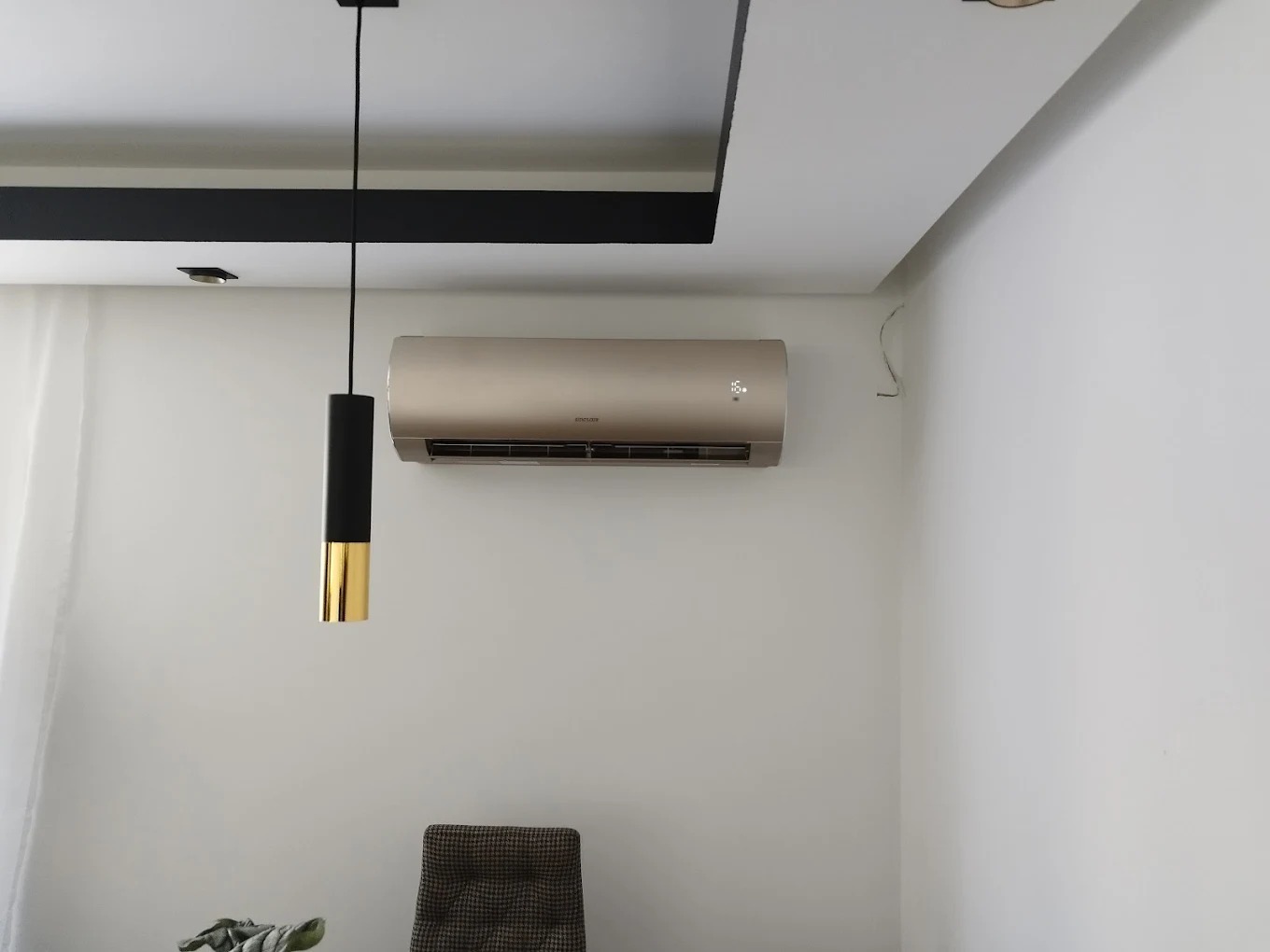 Klimatyzator w jasnym wnętrzu z lampą wiszącą. Widoczne elementy dekoracyjne sufitu. Minimalistyczny design, nowoczesne wykończenie. Kabel wystający ze ściany.