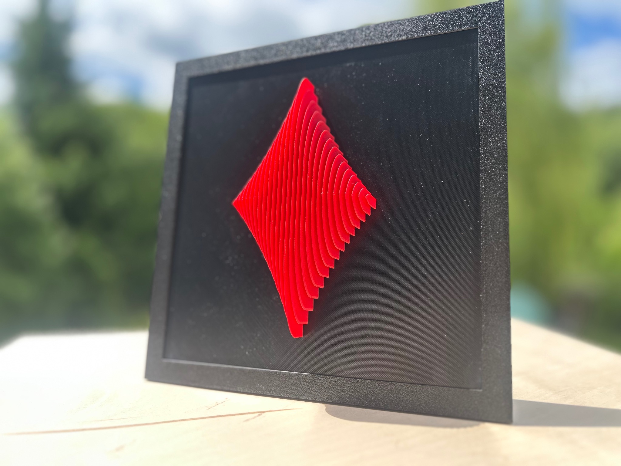 Czarno-czerwony obraz 3D w ramce prezentuje przestrzenny symbol karo, z wyraźną fakturą druku na czarnym tle i rozmyte tło zieleni.