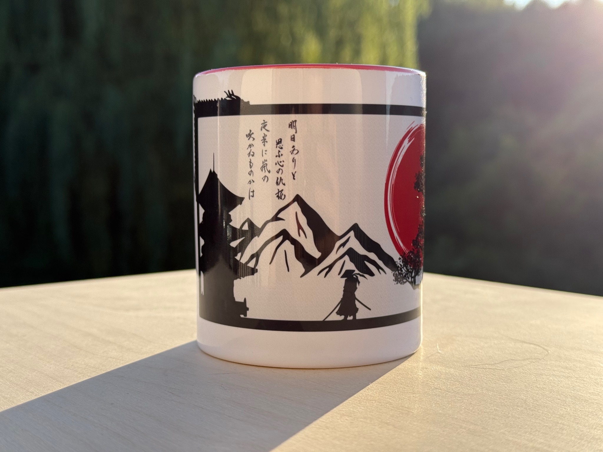 Biały kubek z czarno-czerwonym nadrukiem w stylu japońskim (góry, samuraj, słońce) stoi na drewnianej powierzchni w letnim słońcu.