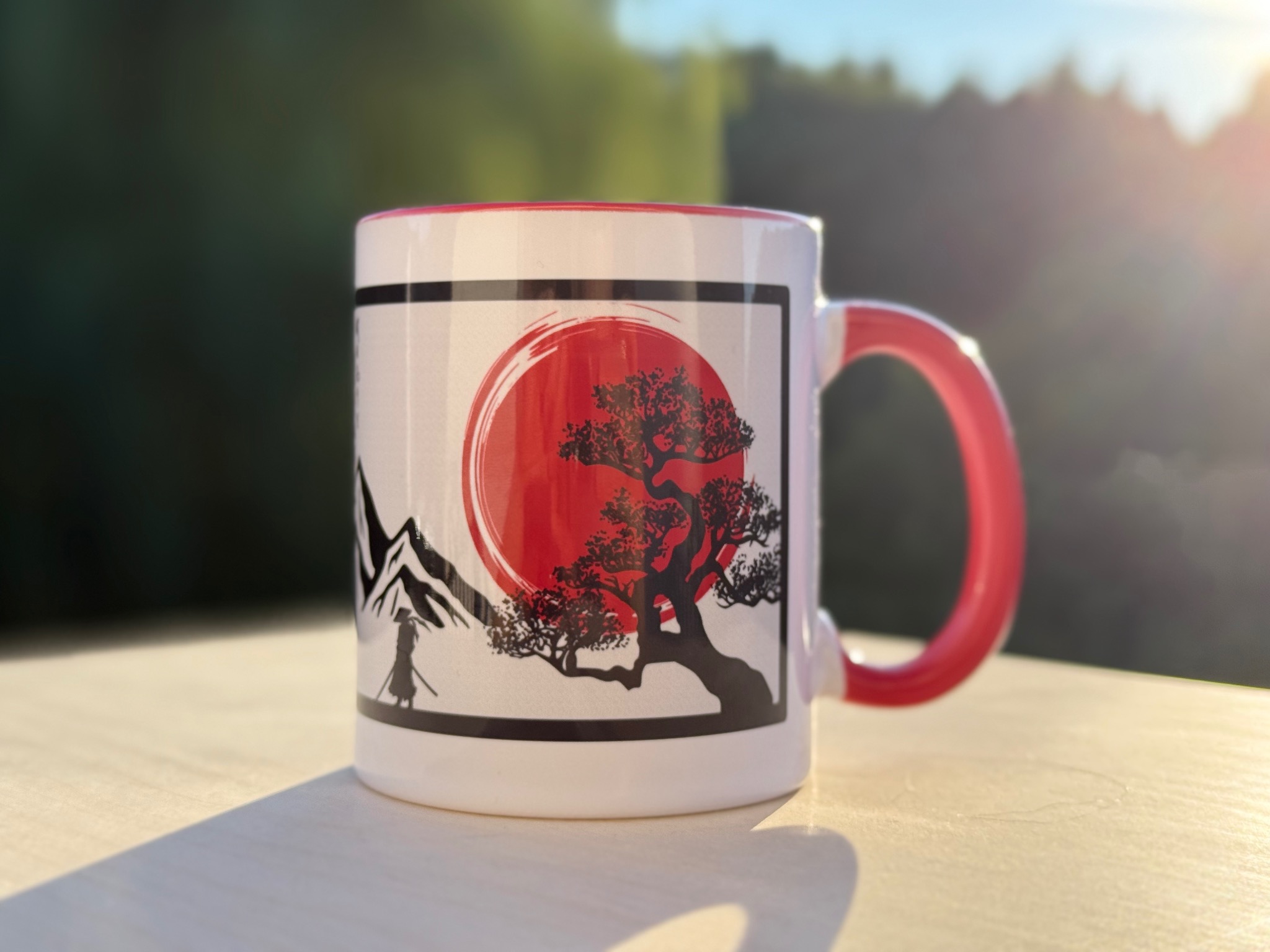 Biały kubek z czerwonym uchem i wnętrzem, na którym widnieje nadruk japońskiego motywu z drzewem, słońcem i sylwetką samuraja na tle zieleni.