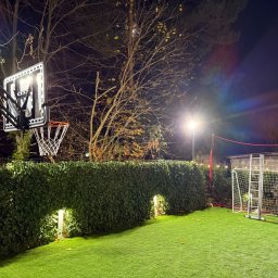Oświetlony, przydomowy plac zabaw nocą: kosz do koszykówki z podświetleniem LED, siatka do siatkówki i bramka do piłki nożnej na sztucznej trawie.
