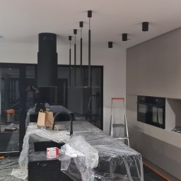 Nowoczesna kuchnia w trakcie montażu oświetlenia: czarne lampy sufitowe i wiszące, drabina, folia ochronna na blacie. Minimalistyczny design, szare szafki.