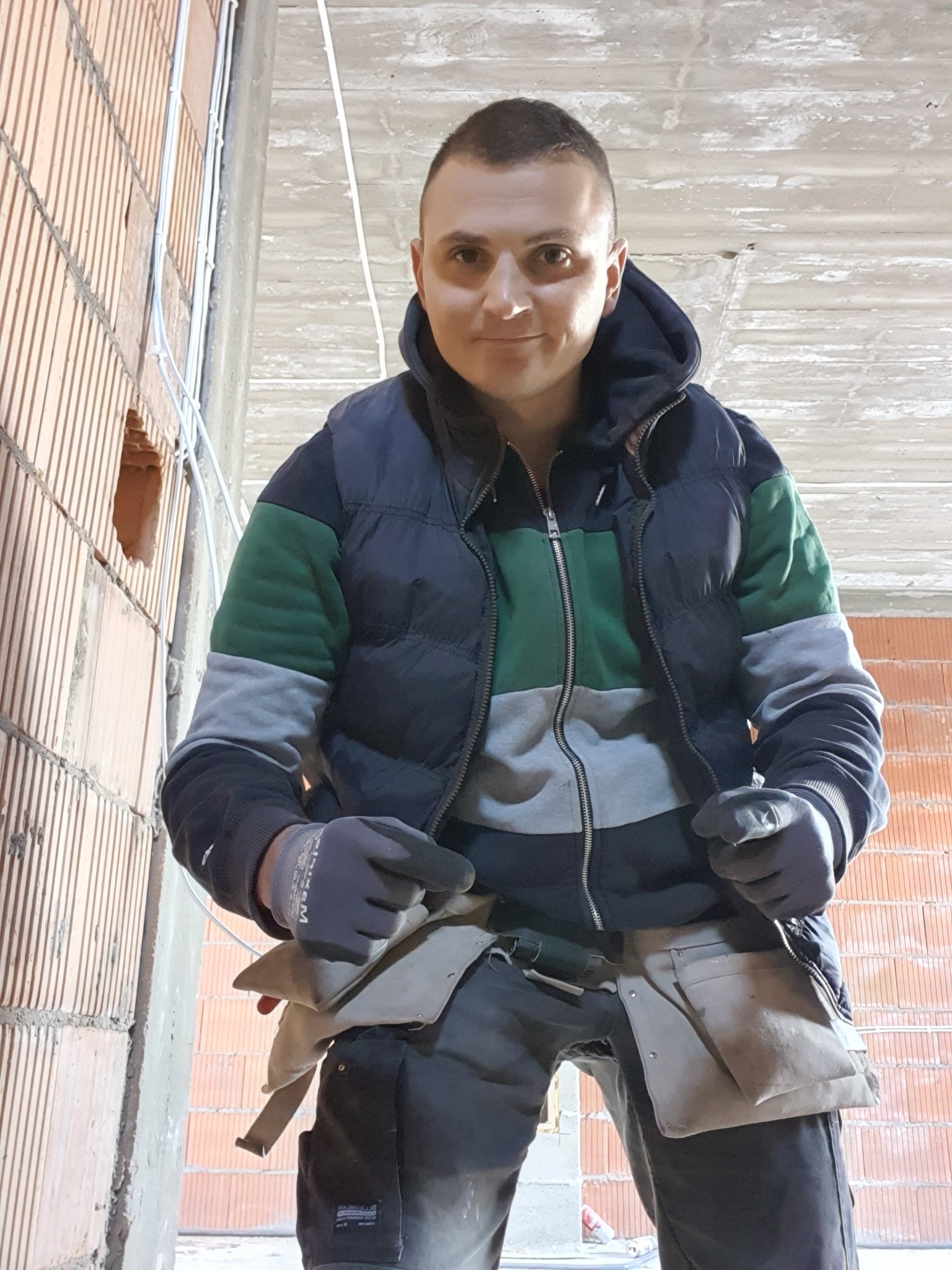 Elektryk w kamizelce i rękawicach, z pasem narzędziowym, prezentuje instalację elektryczną w surowym wnętrzu z ceglanymi ścianami i betonowym sufitem.