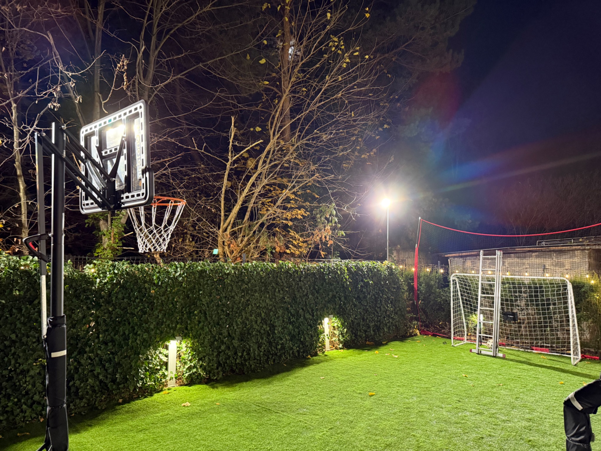 Oświetlony, przydomowy plac zabaw nocą: kosz do koszykówki z podświetleniem LED, siatka do siatkówki i bramka do piłki nożnej na sztucznej trawie.