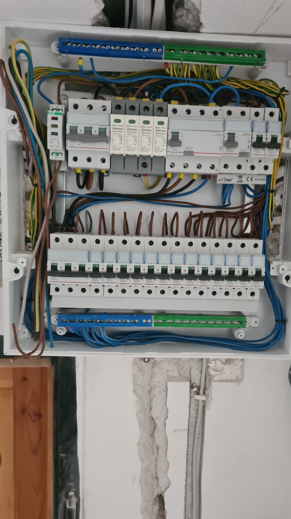 Rozdzielnica elektryczna w białej obudowie z widocznymi bezpiecznikami, przewodami i listwami zaciskowymi. Instalacja w trakcie prac, widoczne ślady po kuciu w ścianie.