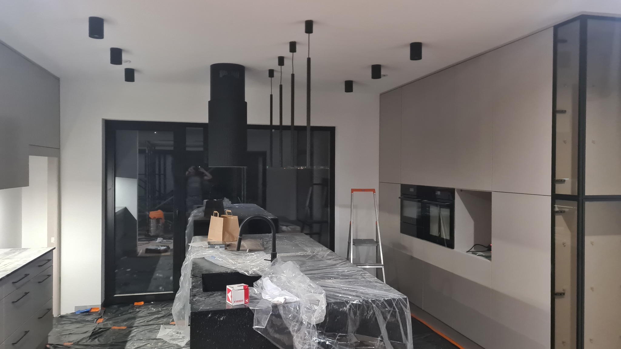 Nowoczesna kuchnia w trakcie montażu oświetlenia: czarne lampy sufitowe i wiszące, drabina, folia ochronna na blacie. Minimalistyczny design, szare szafki.