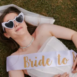Panna młoda leży na trawie w okularach w kształcie serc i z szarfą 'Bride to Be'. Uśmiechnięta kobieta w bieli, z welonem, w słoneczny dzień. Sesja narzeczeńska w plenerze.