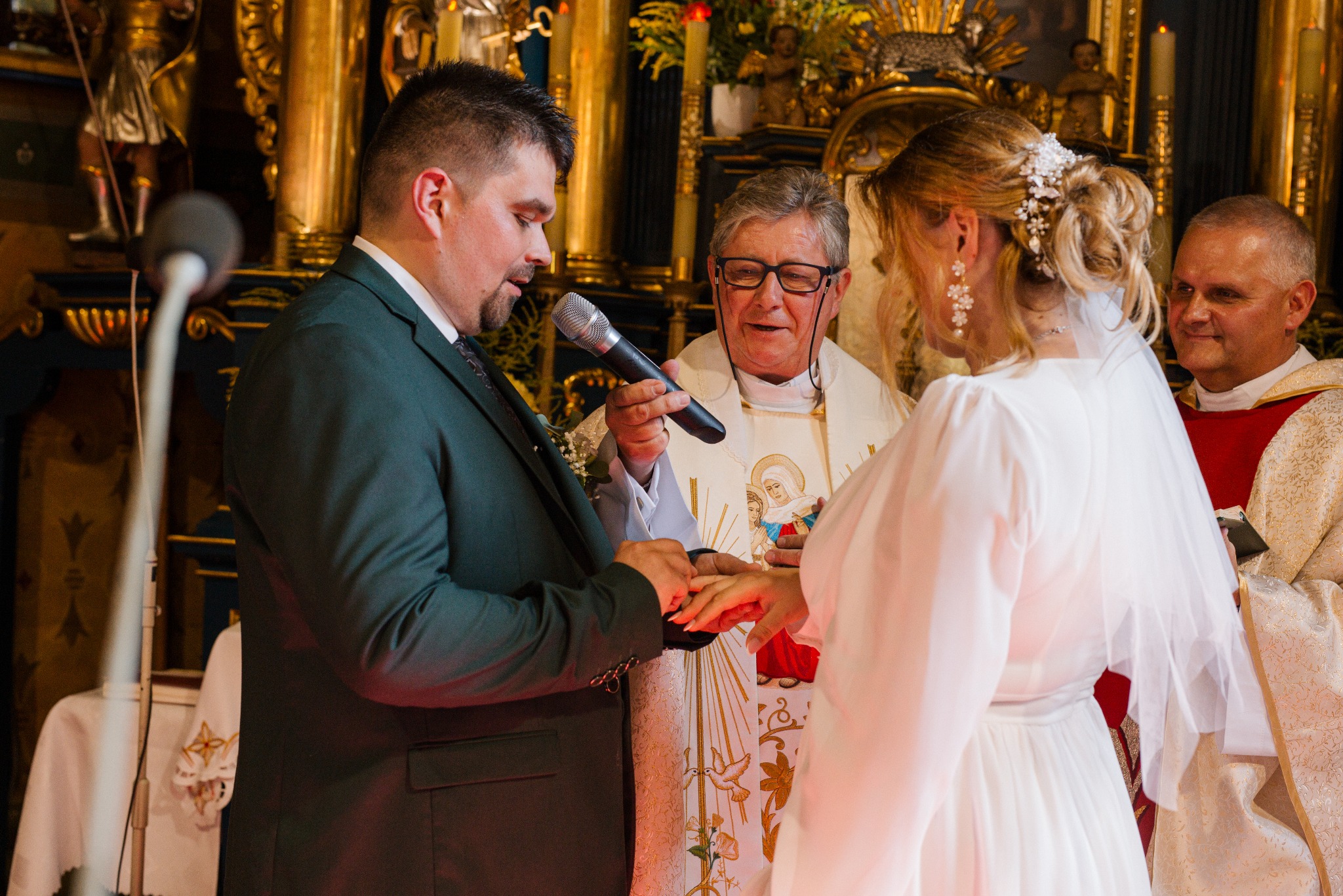 Wymiana obrączek podczas ceremonii ślubnej w kościele. Pan młody zakłada obrączkę pannie młodej, błogosławieństwo udziela ksiądz z mikrofonem. W tle złote zdobienia.