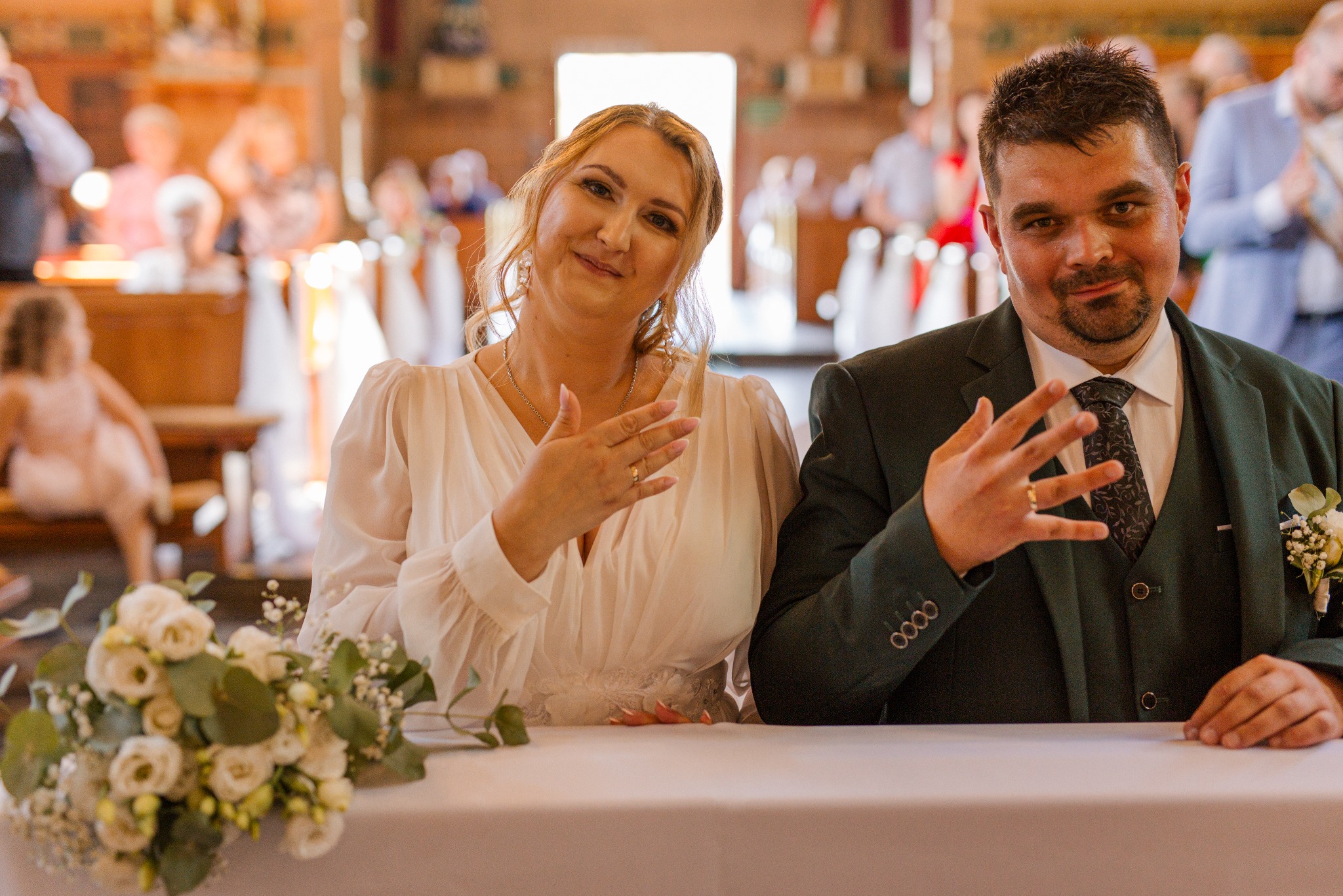 Uśmiechnięta para młoda w kościele, ona w białej sukni z bukietem, on w zielonym garniturze, w tle goście i drewniane ławki. Ujęcie z bliska.