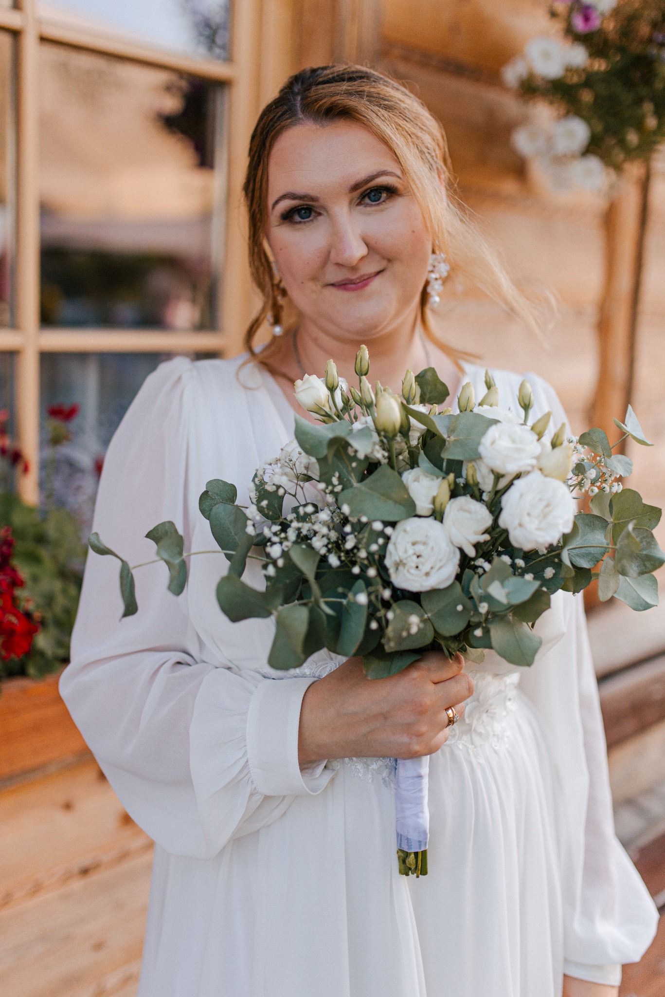 Uśmiechnięta panna młoda w białej sukni trzyma bukiet z białych róż i eukaliptusa na tle drewnianego domu z kwiatami w donicach.
