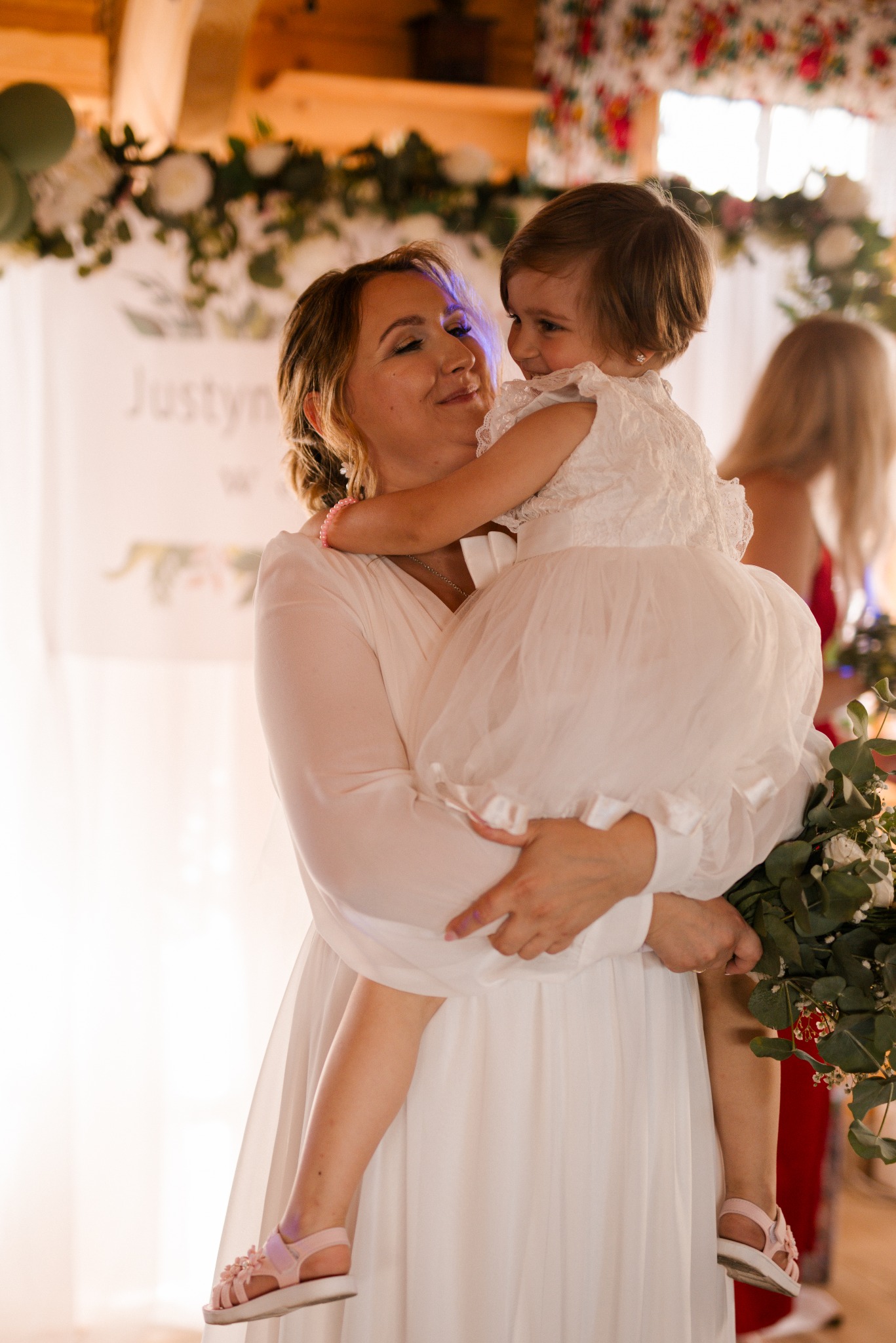 Uśmiechnięta kobieta w białej sukni trzyma na rękach dziewczynkę w białej sukience, w tle dekoracje kwiatowe i osoba w czerwonej sukience.