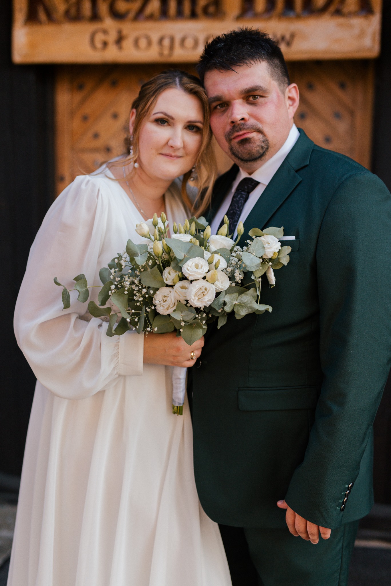 Para młoda: panna młoda w białej sukni trzyma bukiet, pan młody w zielonym garniturze, na tle drewnianych drzwi z napisem 'Karczma Głogów'.
