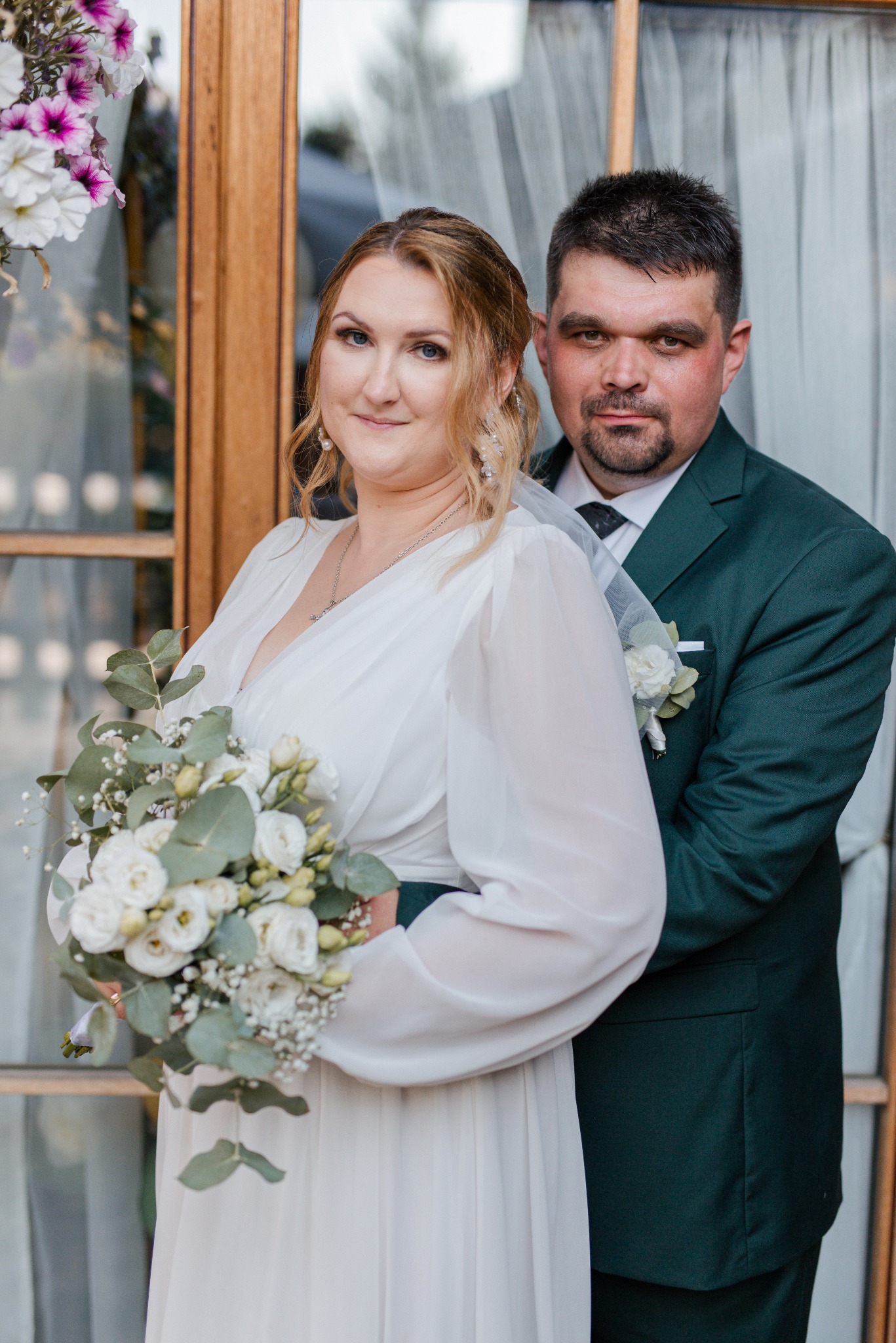 Uśmiechnięta para młoda w objęciach na tle okna: panna młoda w białej sukni z bukietem, pan młody w zielonym garniturze. Romantyczna sesja ślubna.