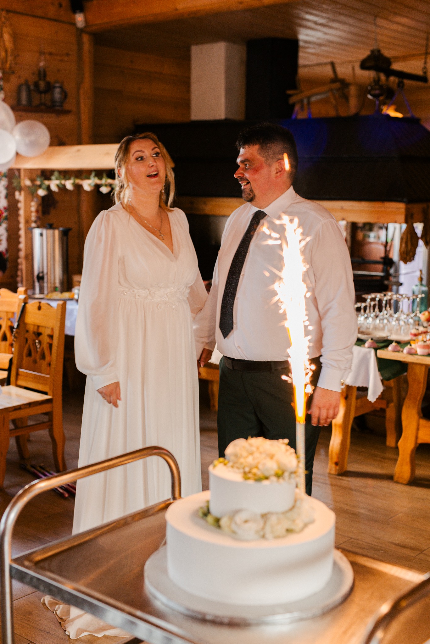 Uśmiechnięta para w rustykalnym wnętrzu, obok dwupiętrowy tort weselny z fontanną iskier, jasna suknia panny młodej i ciemny krawat pana młodego.