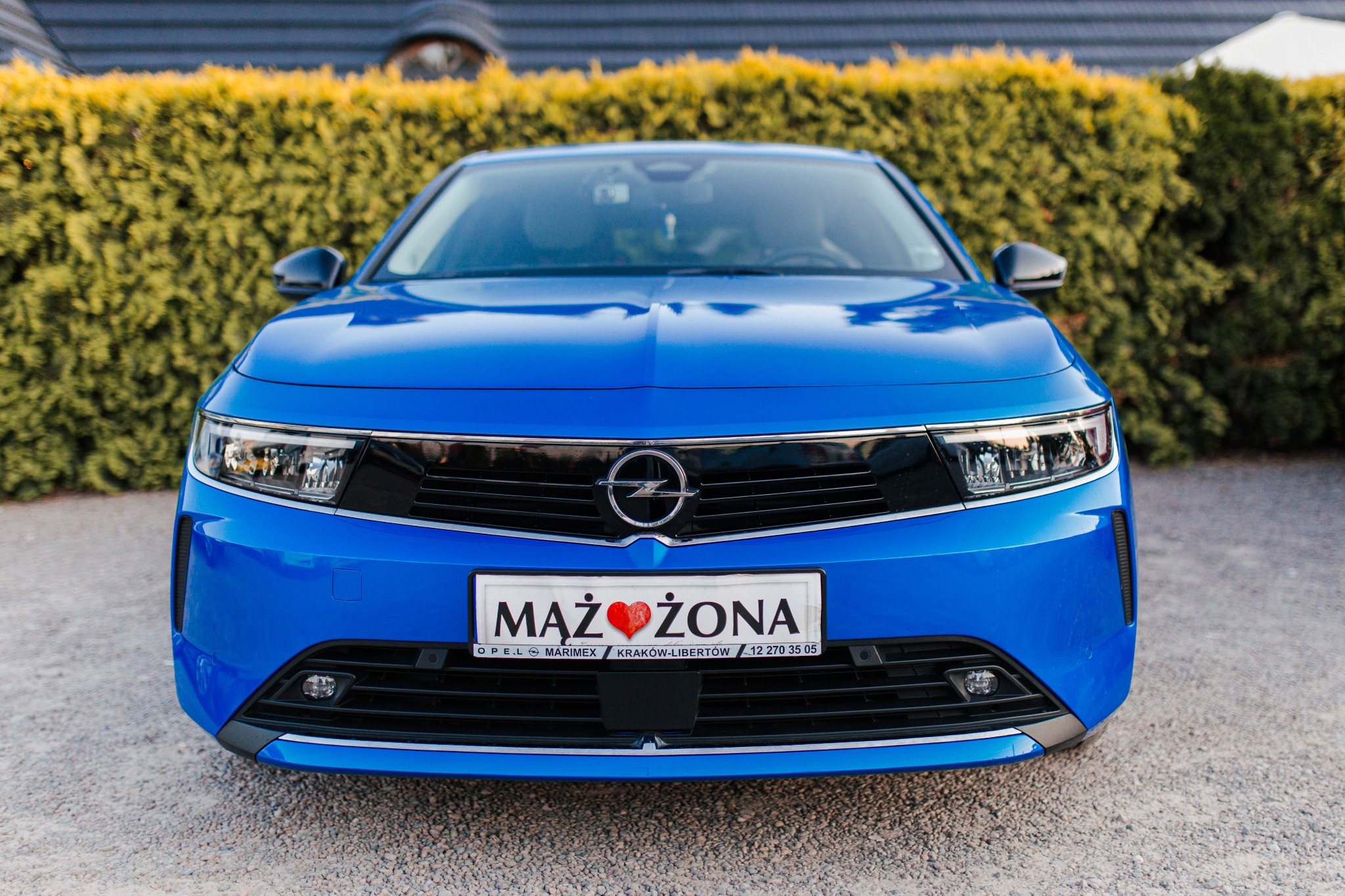 Niebieski Opel Astra z tablicą rejestracyjną 'Mąż Żona' na tle zielonego żywopłotu i szarej elewacji, centralny kadr.