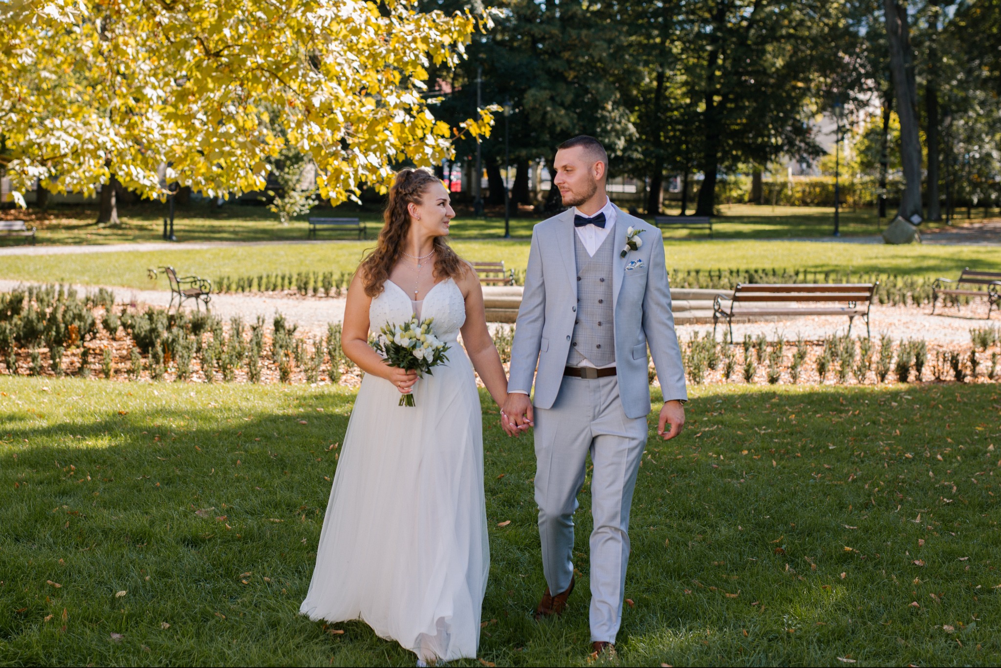 Para młoda w jasnych strojach spaceruje po parku w słoneczny dzień. Panna młoda trzyma bukiet, a drzewa w tle mają jesienne, żółte liście.