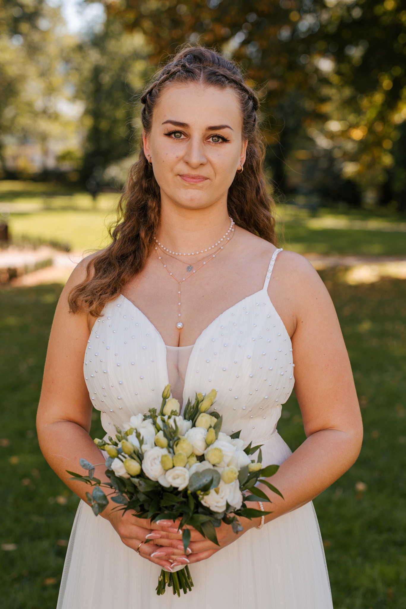 Panna młoda w białej sukni z bukietem w rękach, w plenerze. Delikatny makijaż, perłowy naszyjnik. Ujęcie portretowe, naturalne światło.