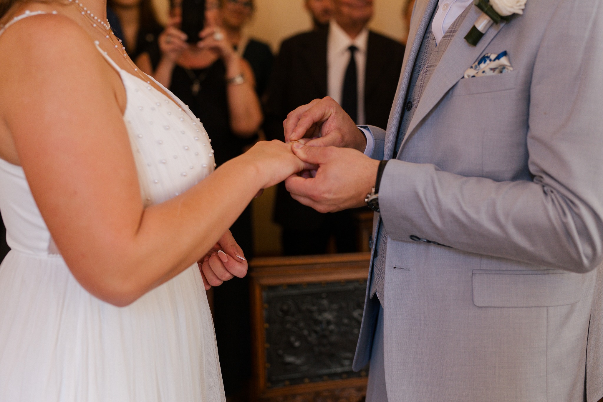 Moment założenia obrączki podczas ceremonii ślubnej. Panna młoda w białej sukni, pan młody w szarym garniturze. W tle goście.