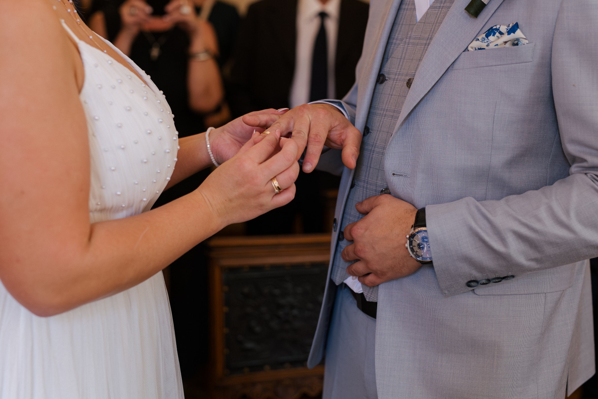 Wymiana obrączek podczas ceremonii ślubnej. Panna młoda w białej sukni i pan młody w popielatym garniturze, zbliżenie na dłonie w trakcie zakładania obrączki.