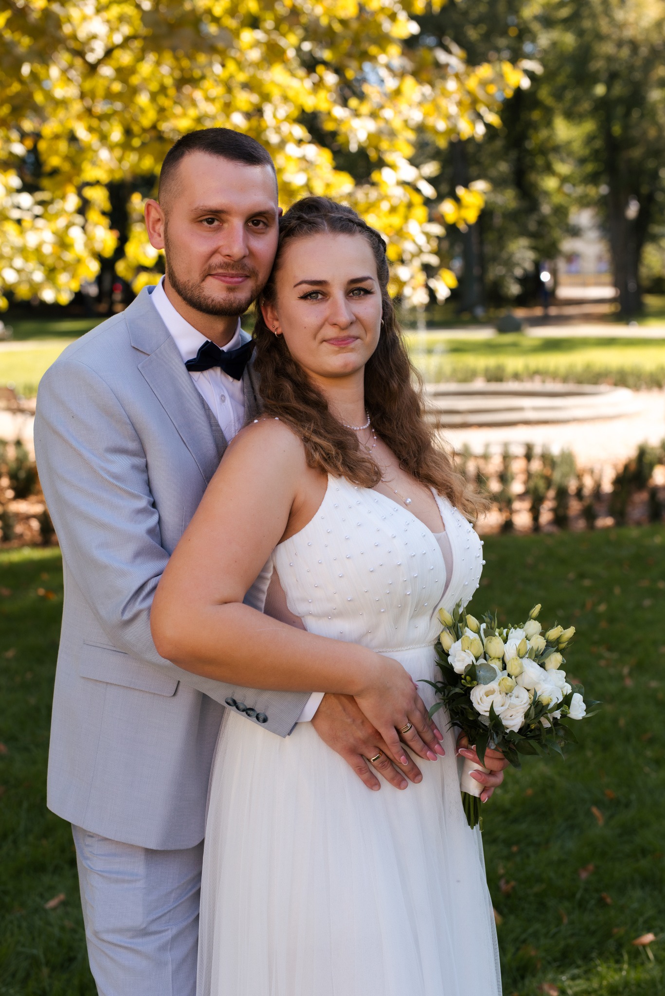 Para młoda w parku: on w szarym garniturze i muszce, ona w białej sukni z bukietem. Jesienne drzewa w tle, miękkie światło, romantyczna atmosfera.