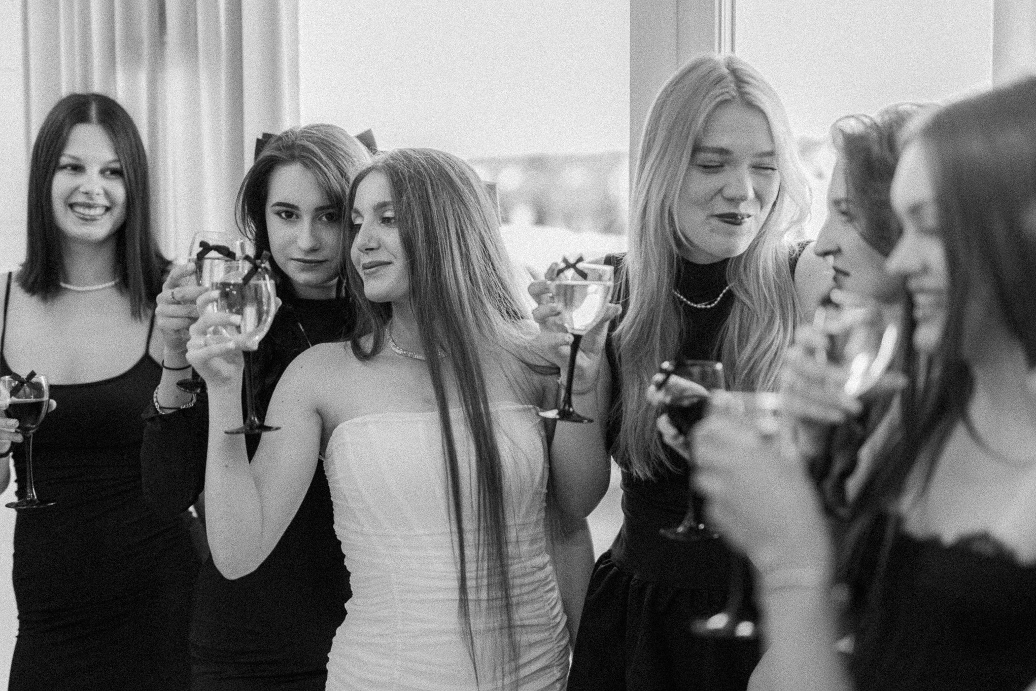 Grupa młodych kobiet w sukienkach, w czerni i bieli, wznosi toast kieliszkami wina. Uśmiechy, elegancja i celebracja w minimalistycznym stylu.