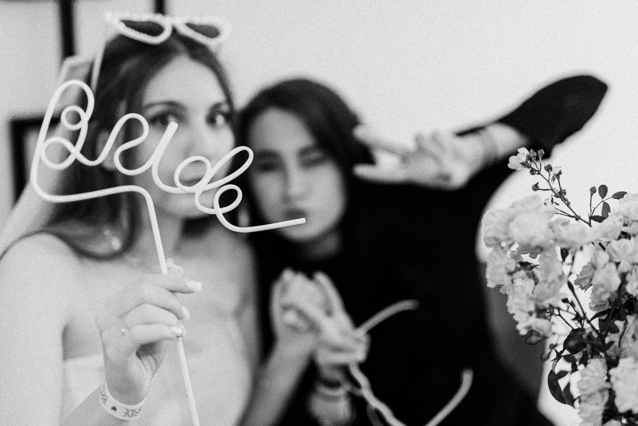 Czarno-białe zdjęcie dwóch kobiet, jedna trzyma rekwizyt z napisem 'Bride', kwiaty z boku, uśmiechy, w tle dekoracje. Fotografia ślubna.