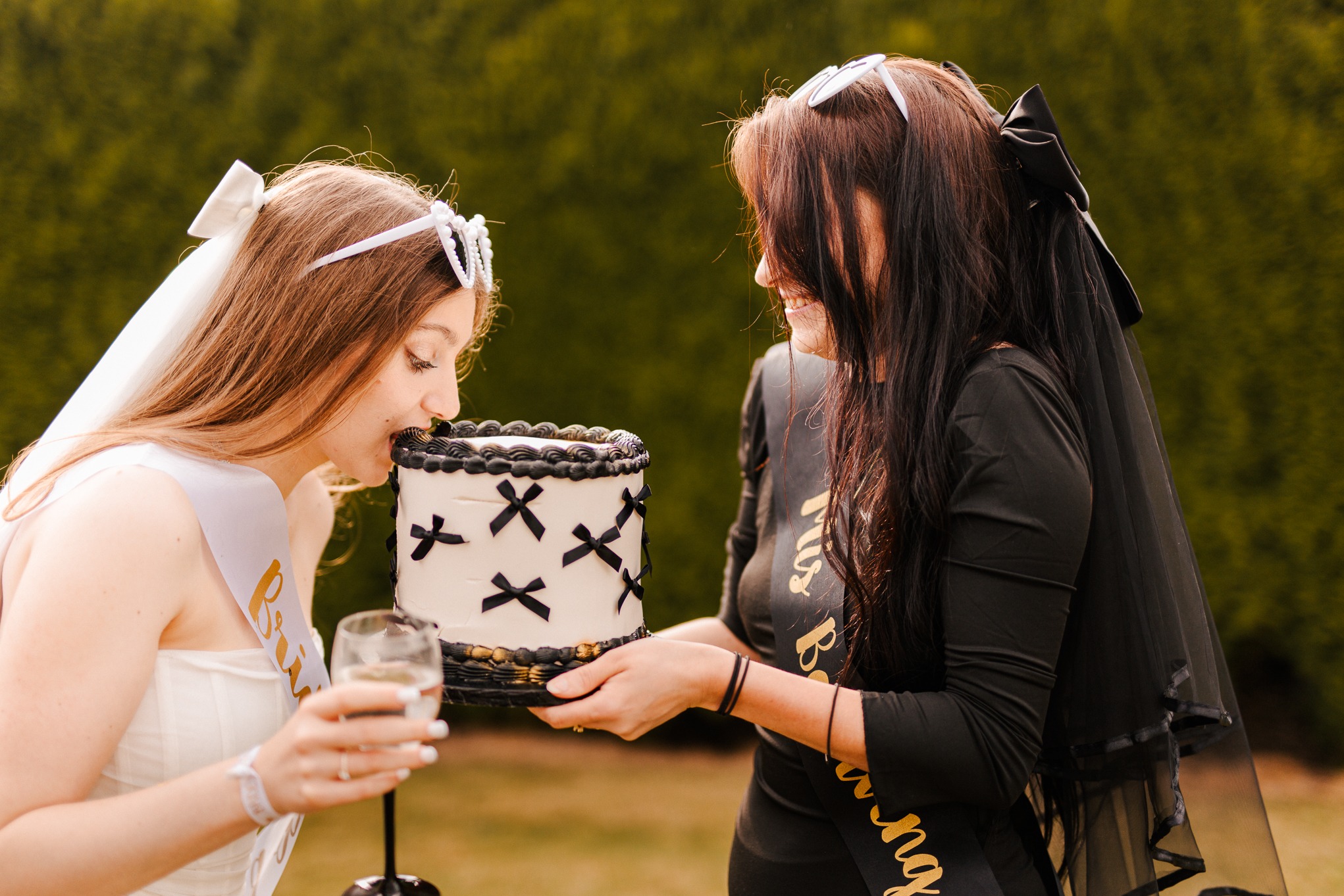 Dwie kobiety, panna młoda w bieli i druhna w czerni, dzielą się tortem podczas wieczoru panieńskiego. Panna młoda je tort, trzymając kieliszek wina.