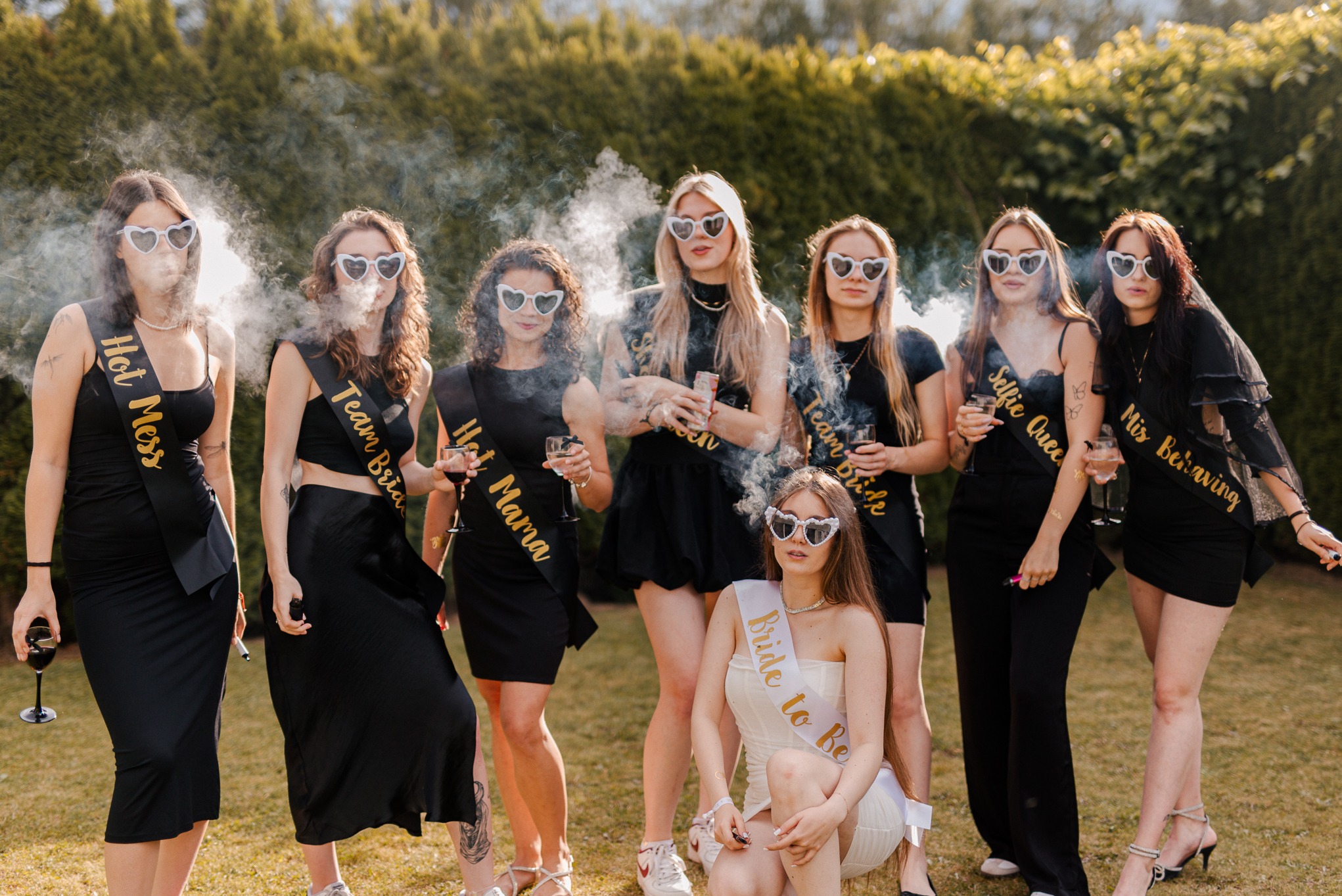 Wieczór panieński: grupa kobiet w okularach w kształcie serc i szarfach, z dymem i drinkami na tle zieleni. Jedna z kobiet kuca na pierwszym planie.